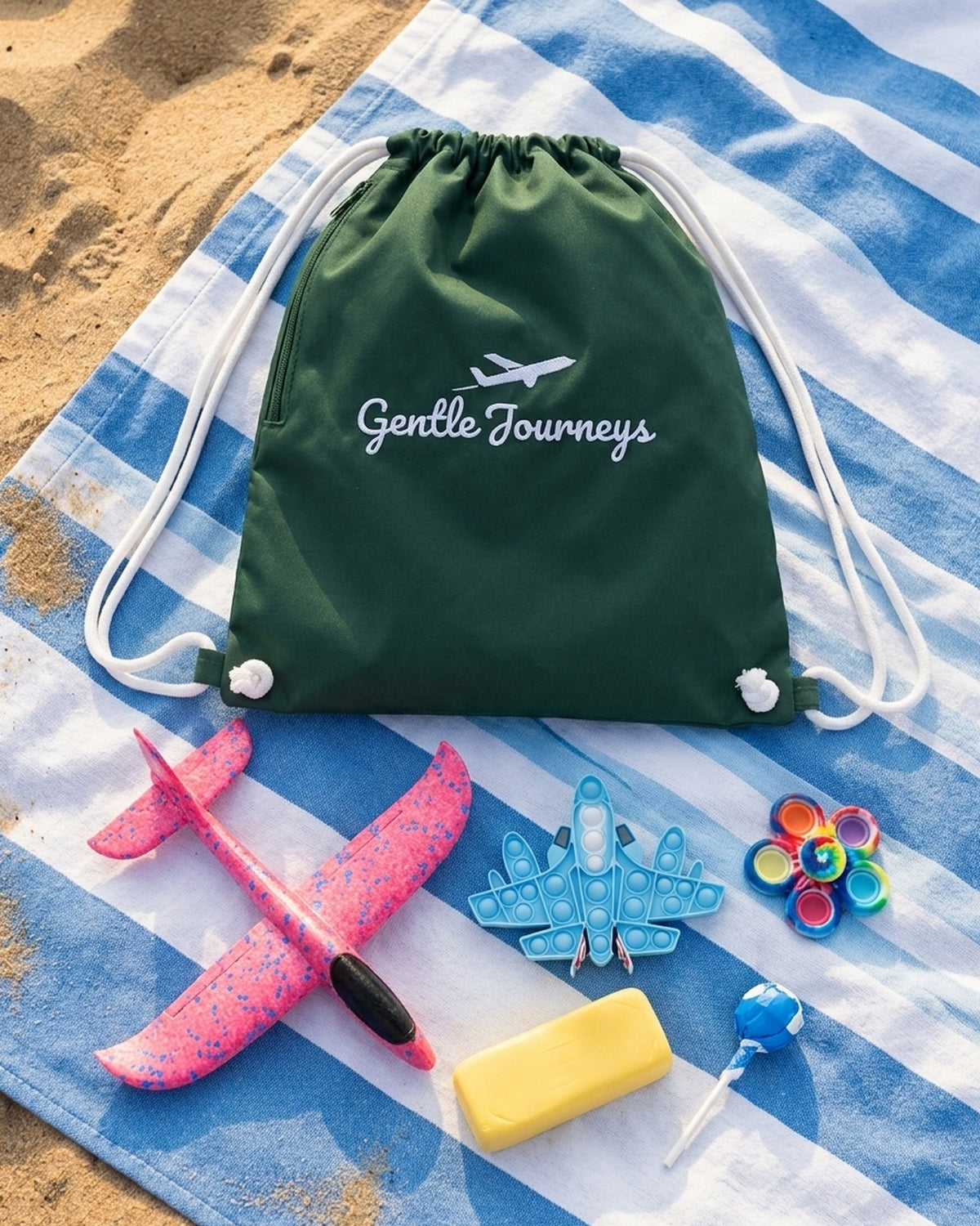 Green Water-Resistant sensory travel bag on a striped beach towel with the foam glider, aeroplane pop it, tie-dye spinner, butter stress bar and sugar-free lollipop